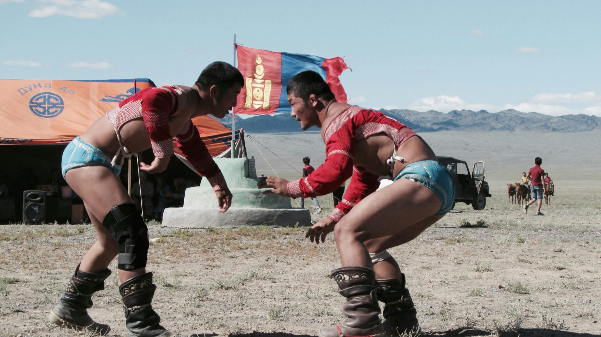Two men compete in traditional Mongolian wrestling at a festival outdoors with cultural elements visible.